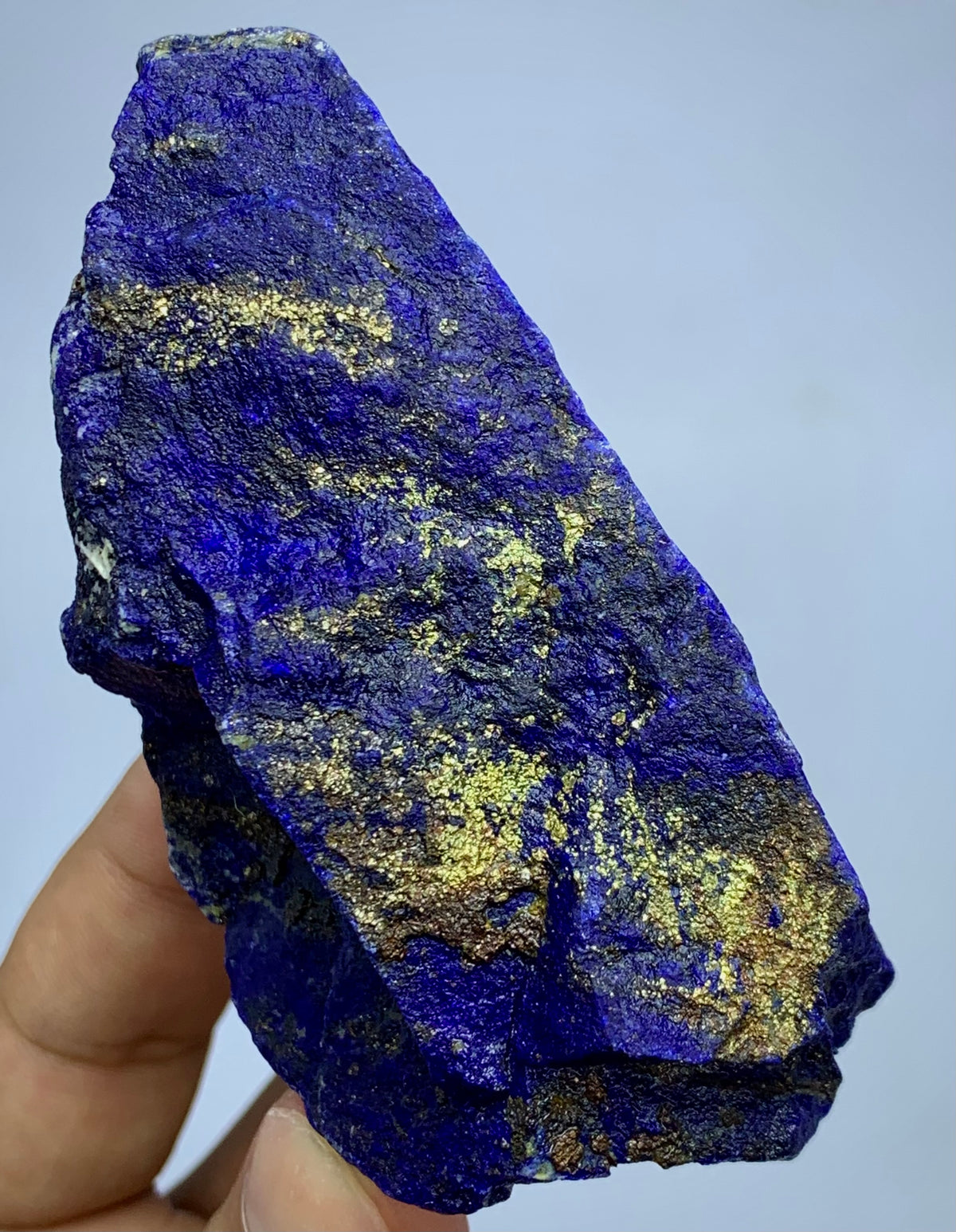 A raw, rough-cut lapis lazuli crystal held in a person's hand against a light blue-gray background. The crystal is deep blue with prominent metallic gold pyrite inclusions.
