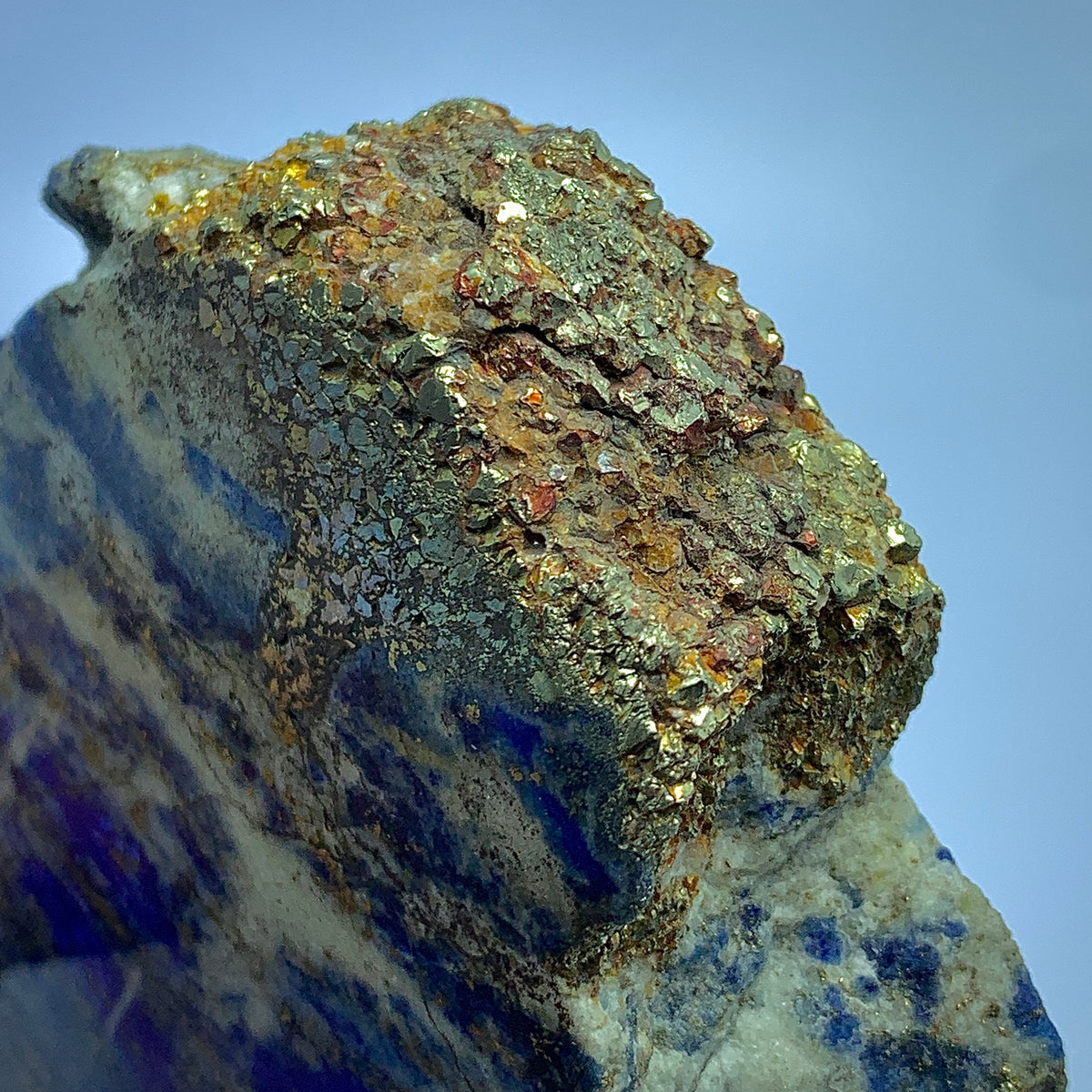 A close-up of a rough mineral specimen with metallic gold, copper, and dark gray crystalline texture on a pale blue and white base, set against a clear light blue sky.