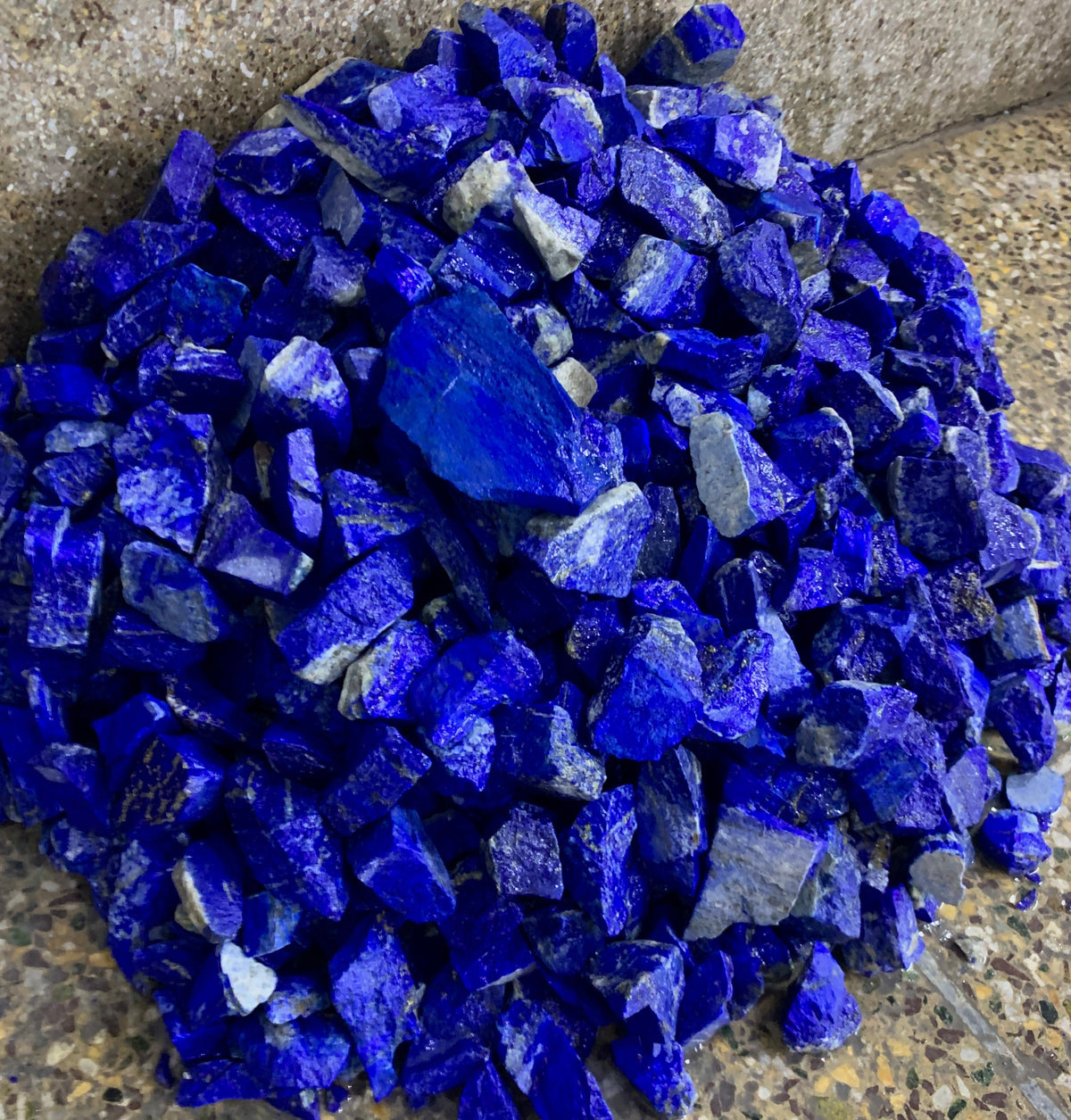 A pile of raw, rough-cut cobalt blue lapis lazuli crystal chips resting on a speckled stone floor.