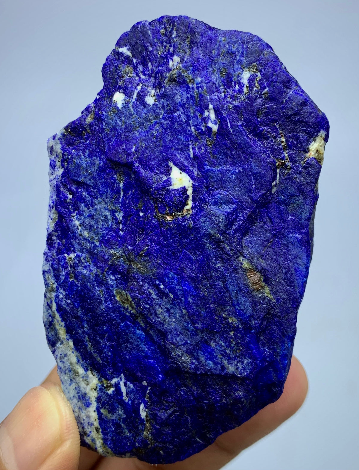 A raw, rough-cut lapis lazuli crystal specimen held in a hand, showcasing its deep blue color with natural white and gold matrix inclusions, against a light gray background.