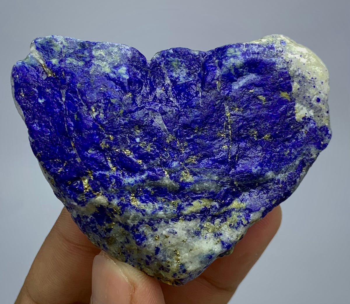 A heart-shaped lapis lazuli crystal specimen with deep blue and purple coloration and gold pyrite flecks, held in a person's hand against a neutral gray background.