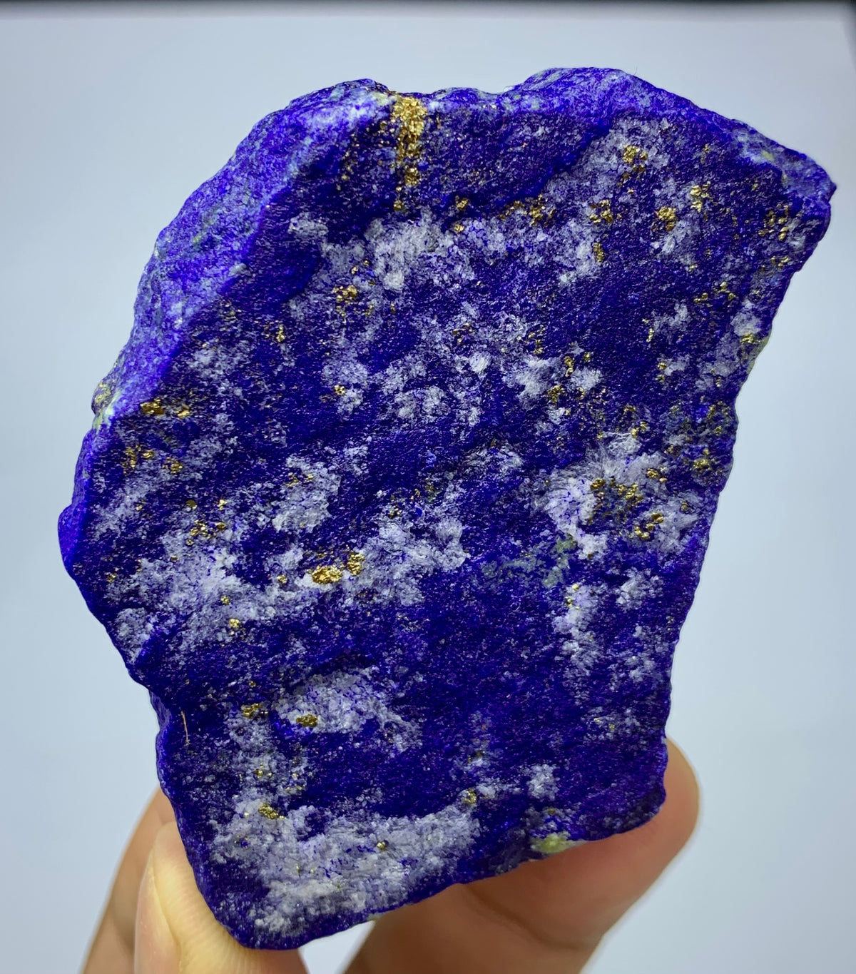 A raw, rough-cut lapis lazuli crystal specimen held in a hand, showcasing its deep blue-violet color with natural gold and white inclusions.