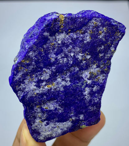A raw, rough-cut lapis lazuli crystal specimen held in a hand, showcasing its deep blue-violet color with natural gold and white inclusions.