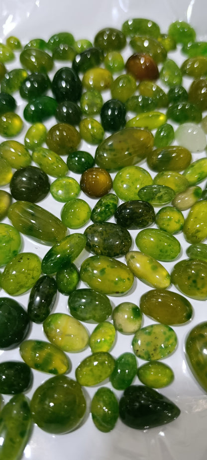 A pile of polished green gemstone beads in various oval and teardrop shapes, showing shades of forest green, olive, and yellow-green on a white background.