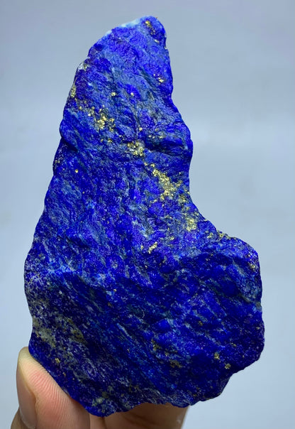 A raw, rough Lapis Lazuli crystal specimen held by a hand, showcasing its deep blue color with natural gold pyrite inclusions against a neutral gray background.