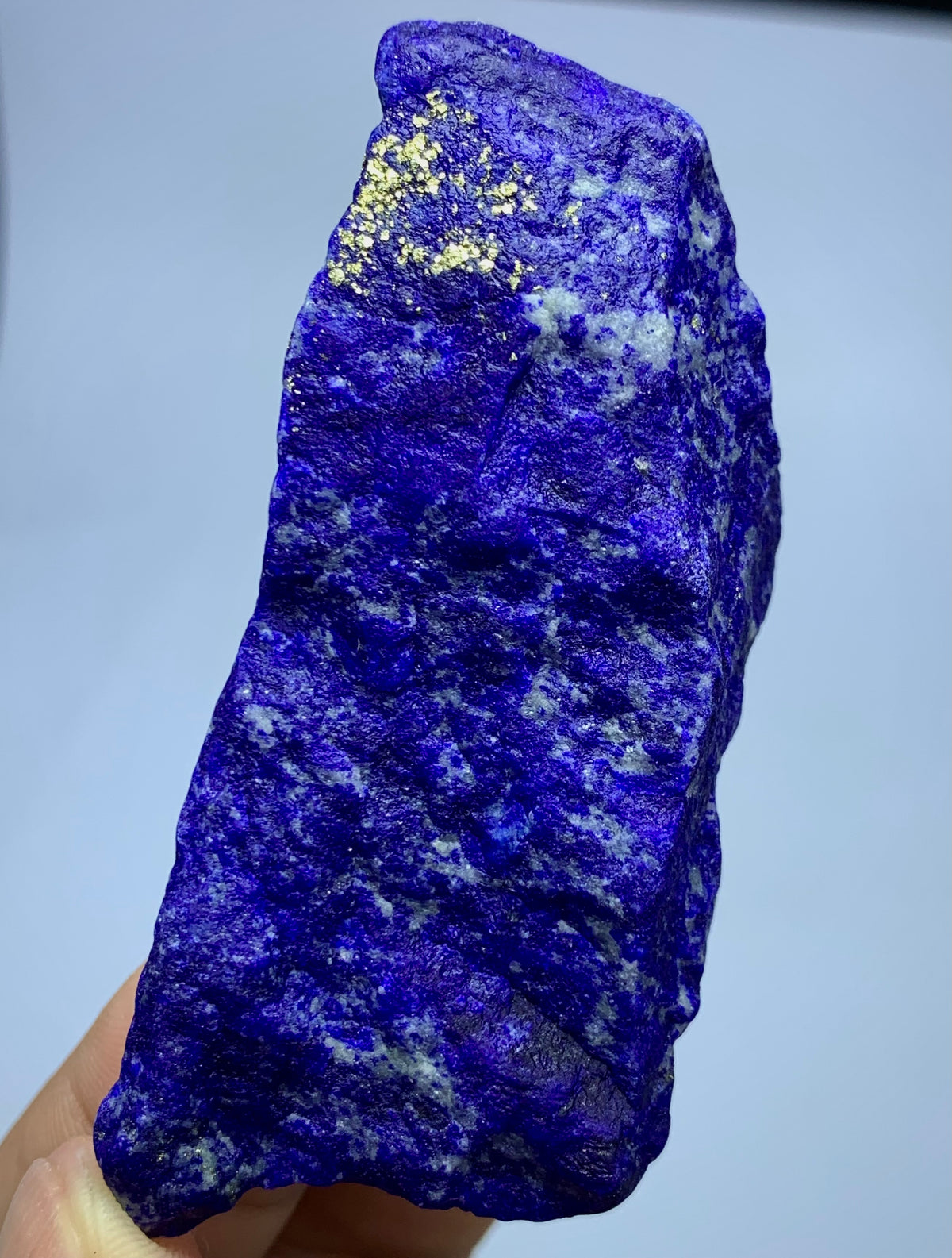 A raw, rough-cut lapis lazuli crystal specimen held in a hand, showcasing its deep blue and purple coloration with natural white and gold matrix inclusions.