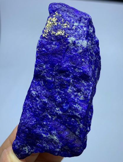 A raw, rough-cut lapis lazuli crystal specimen held in a hand, showcasing its deep blue and purple coloration with natural white and gold matrix inclusions.
