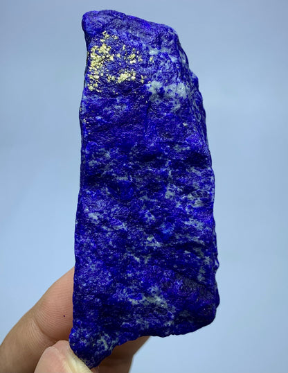 A raw, rough-cut blue lapis lazuli crystal specimen with bright gold pyrite flecks, held in a person's hand against a light blue background.
