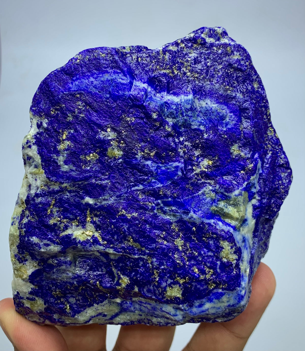 A raw lapis lazuli crystal specimen with deep blue and purple marbling and gold pyrite flecks, held in a person's hand against a light gray background.
Detail shot of Lazurite blue coloring on rough specimen
700g Lapis Lazuli specimen with scale reference
Mine 4 Lapis Lazuli, close-up on Pyrite cluster
