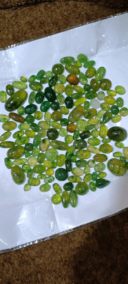 A pile of polished green gemstone chips or beads scattered on a white sheet of paper, photographed from above on a brown textured surface.