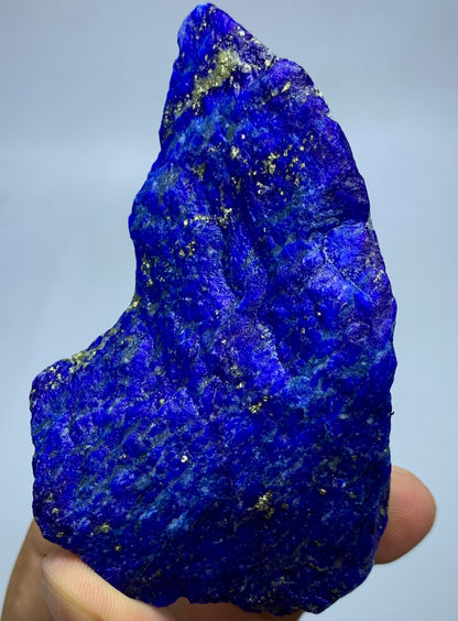 A raw, rough-cut blue lapis lazuli crystal specimen with visible gold pyrite inclusions, held in a person's hand against a light gray background.