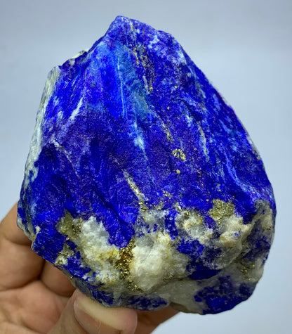 A hand holding a raw lapis lazuli crystal specimen, showing deep blue coloration with white matrix and metallic gold pyrite inclusions against a plain light background.