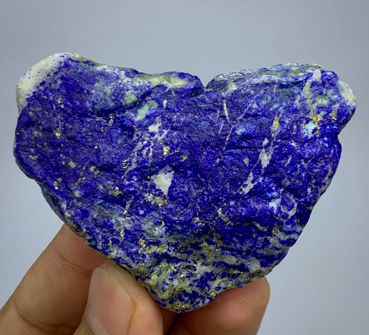 A heart-shaped raw lapis lazuli crystal held in a person's hand against a plain light gray background.