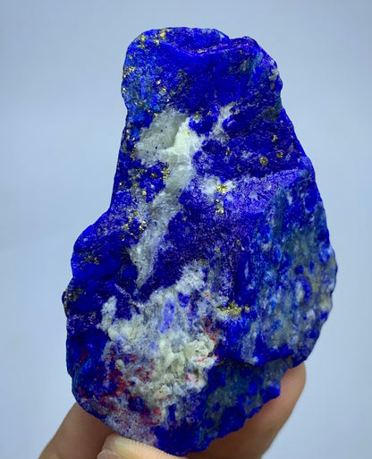 A raw blue lapis lazuli crystal specimen with white and gold mineral inclusions, held in a person's hand against a light gray background.