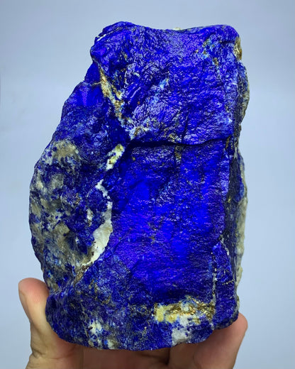 A raw lapis lazuli crystal specimen with deep blue coloration and gold pyrite veins, held in a hand against a light gray background.