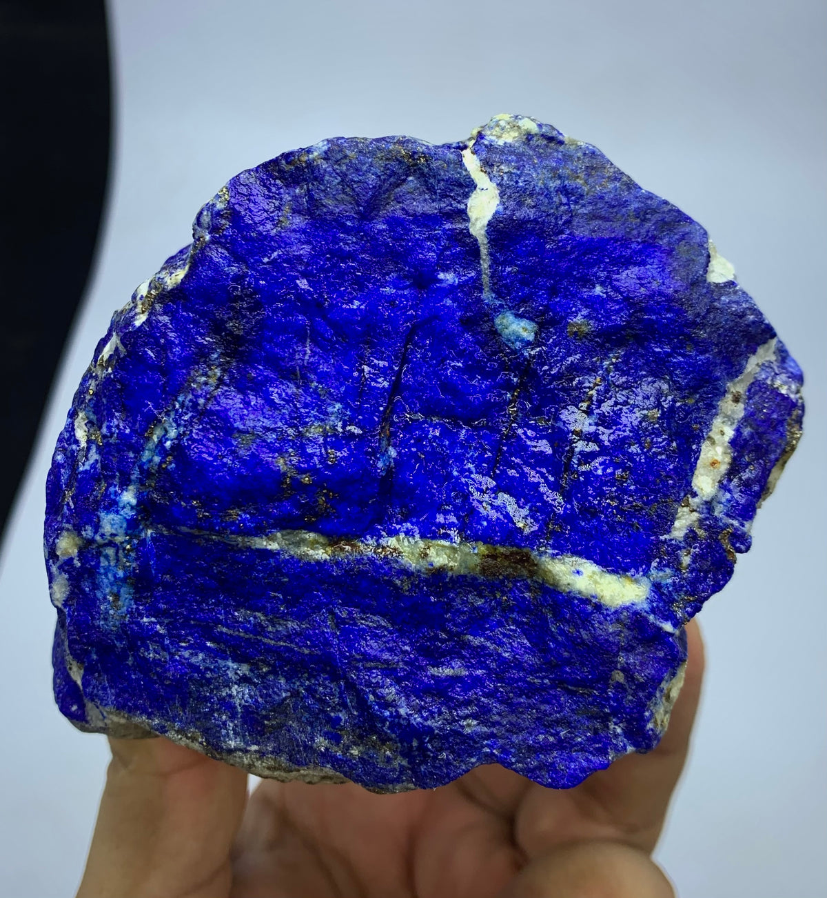 A raw lapis lazuli crystal specimen with deep blue coloration and white veins, held in a person's hand against a plain light background.