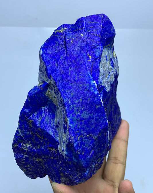 A raw, rough blue lapis lazuli crystal held in a person's hand against a plain light background.