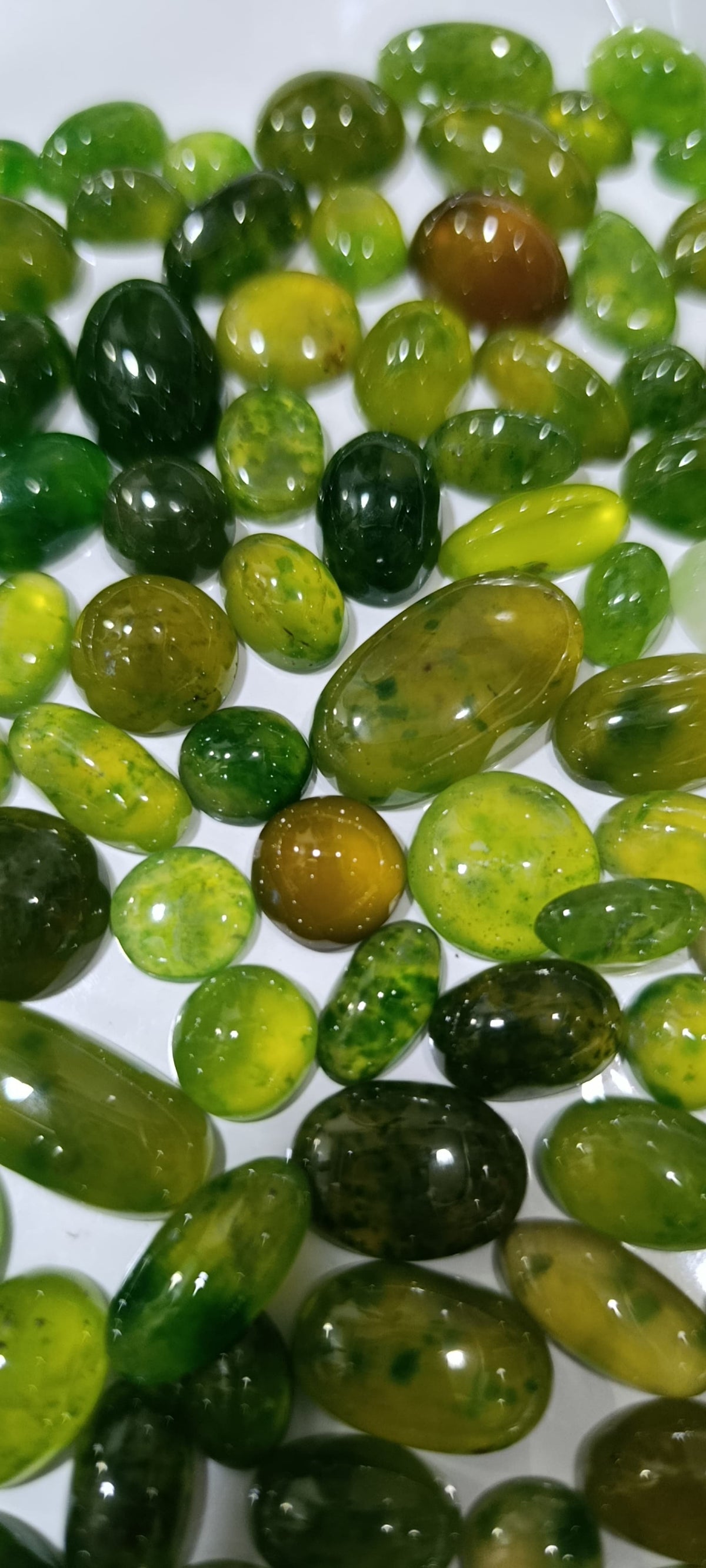A close-up pile of polished green gemstone beads or cabochons in various oval and rounded shapes, showing translucent shades from light yellow-green to deep forest green on a white background.