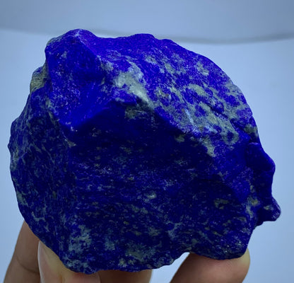 A raw, rough-cut piece of lapis lazuli crystal held in a hand against a light blue background.