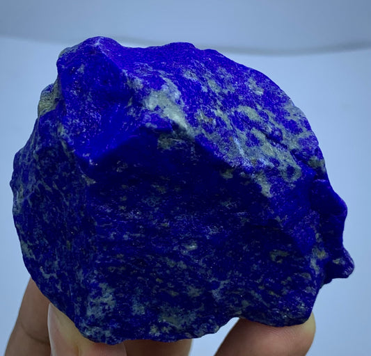 A raw, rough-cut piece of lapis lazuli crystal held in a hand against a light blue background.