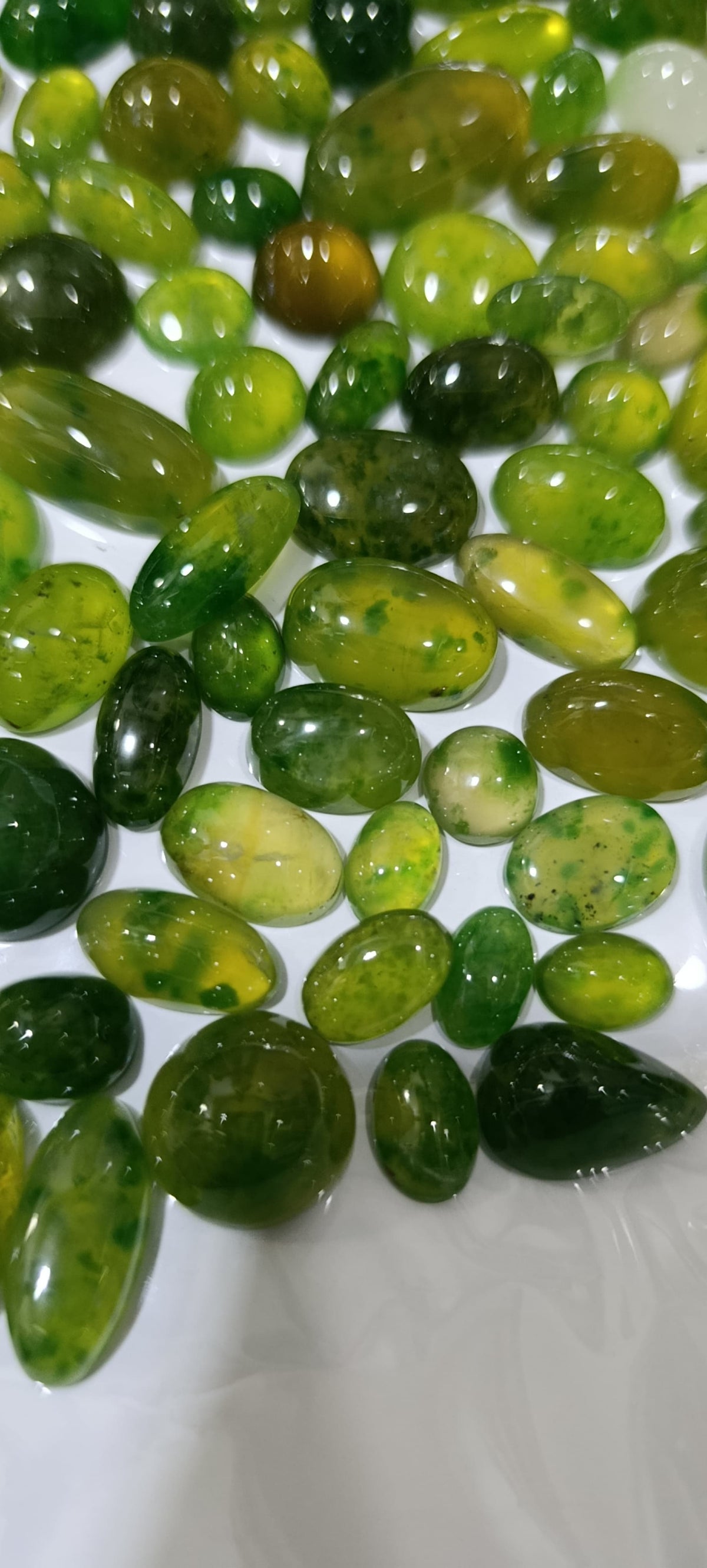 A pile of polished green gemstone cabochons in various oval and teardrop shapes, showing natural marbling and speckles of yellow and lighter green, arranged on a white surface.