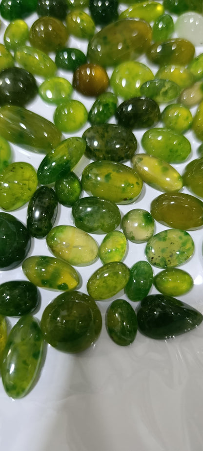 A pile of polished green gemstone cabochons in various oval and teardrop shapes, showing natural marbling and speckles of yellow and lighter green, arranged on a white surface.