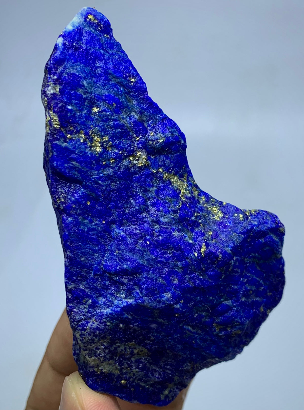 A raw, rough-cut piece of lapis lazuli crystal held up by a hand, showing its deep blue color with natural gold pyrite inclusions against a neutral gray background.
