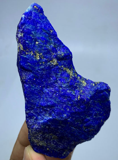 A raw, rough-cut piece of lapis lazuli crystal held up by a hand, showing its deep blue color with natural gold pyrite inclusions against a neutral gray background.