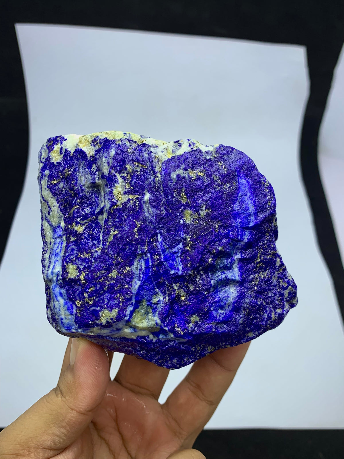A hand holding a rough, unpolished lapis lazuli crystal specimen with deep blue and purple coloration and small gold pyrite inclusions, set against a white sheet of paper background.
Detail shot of Lazurite blue coloring on rough specimen
700g Lapis Lazuli specimen with scale reference