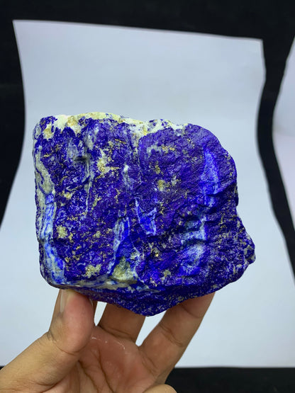 A hand holding a rough, unpolished lapis lazuli crystal specimen with deep blue and purple coloration and small gold pyrite inclusions, set against a white sheet of paper background.
Detail shot of Lazurite blue coloring on rough specimen
700g Lapis Lazuli specimen with scale reference