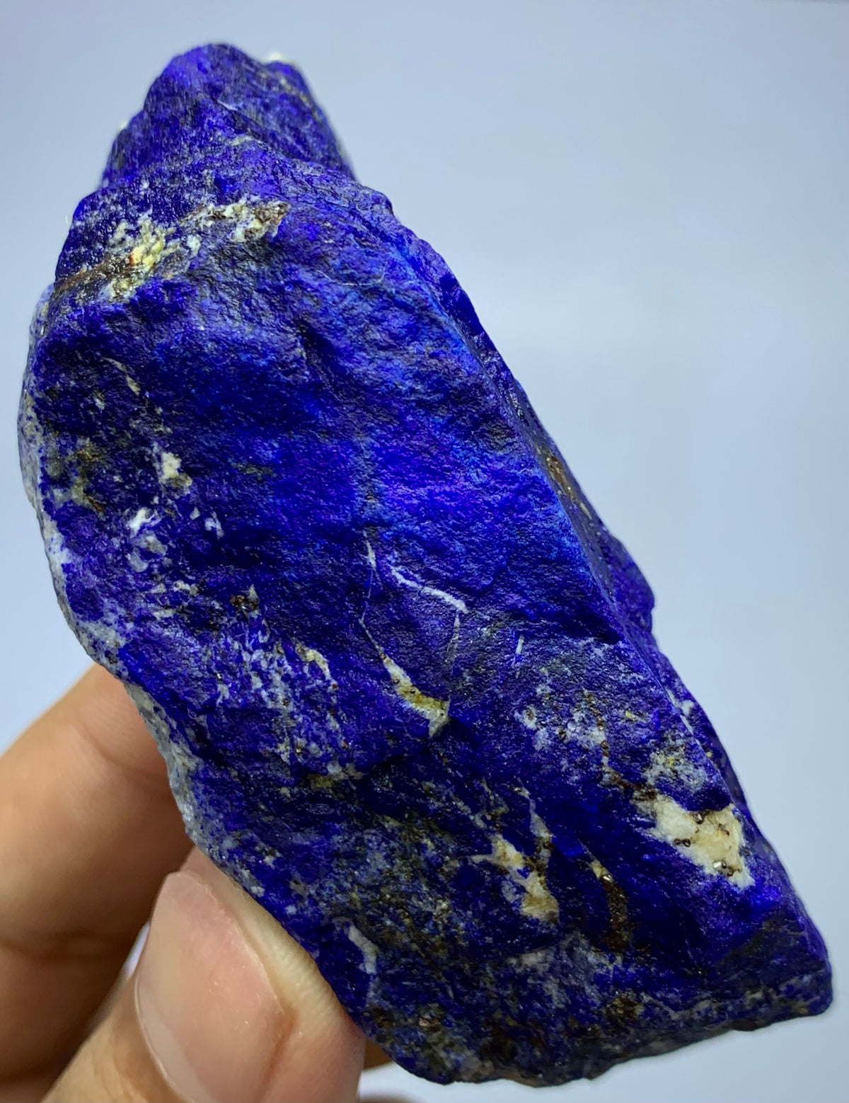A raw, rough-cut piece of lapis lazuli crystal held up by a hand, showing its deep blue color with natural gold and white matrix inclusions.