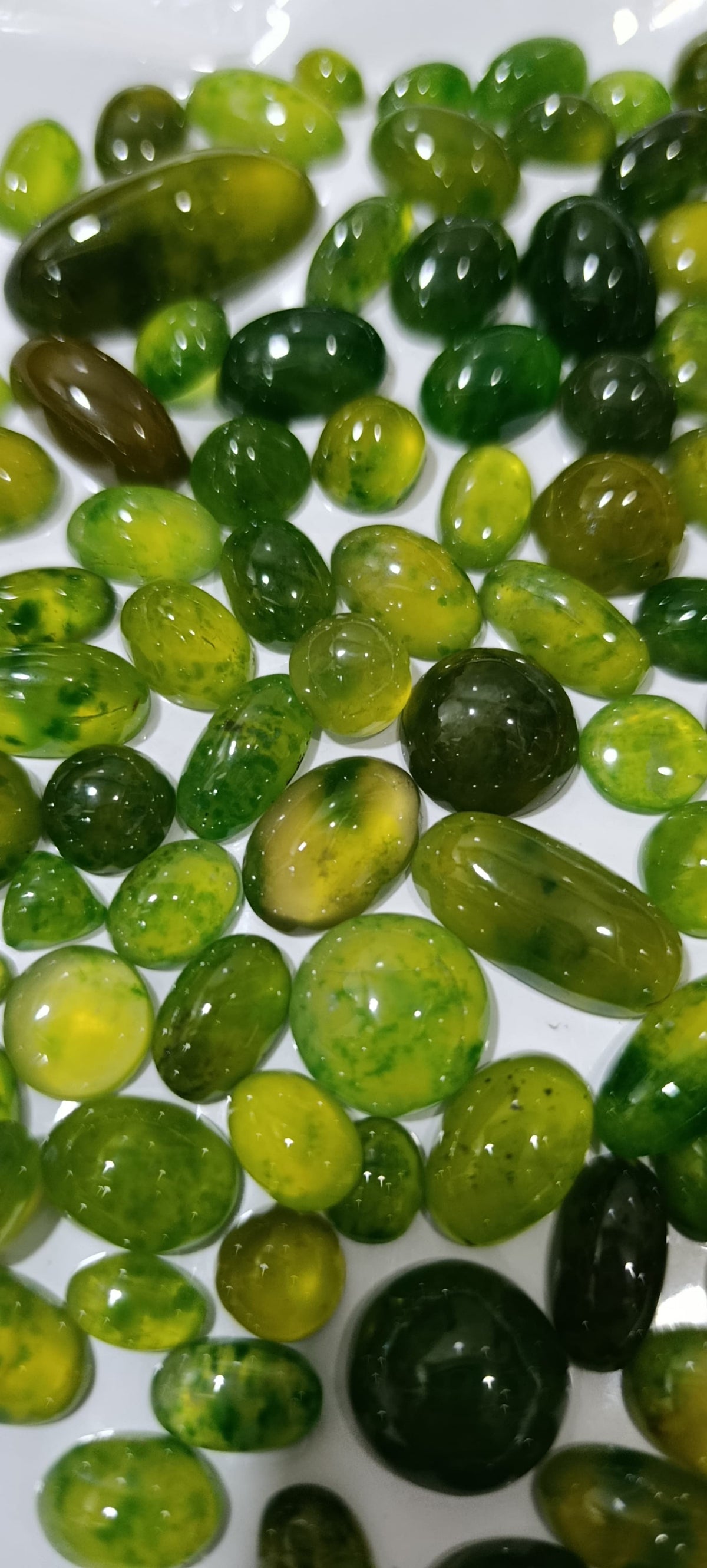 A close-up pile of polished green gemstone beads or cabochons on a white surface, showing translucent shades from light yellow-green to deep forest green.