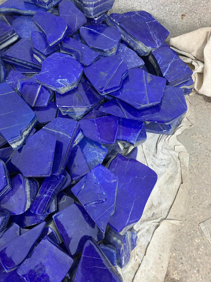 A pile of polished cobalt blue stone chips or tiles with natural rough edges, resting on a light gray sheet of paper on a concrete surface.