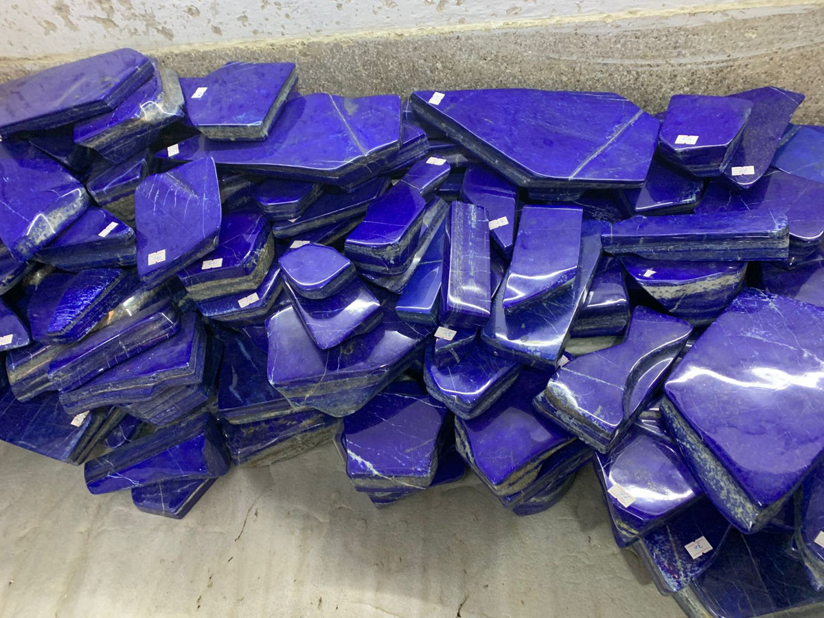 A large pile of polished royal blue lapis lazuli crystal slabs wrapped in protective plastic, stacked on a concrete floor against a concrete wall.