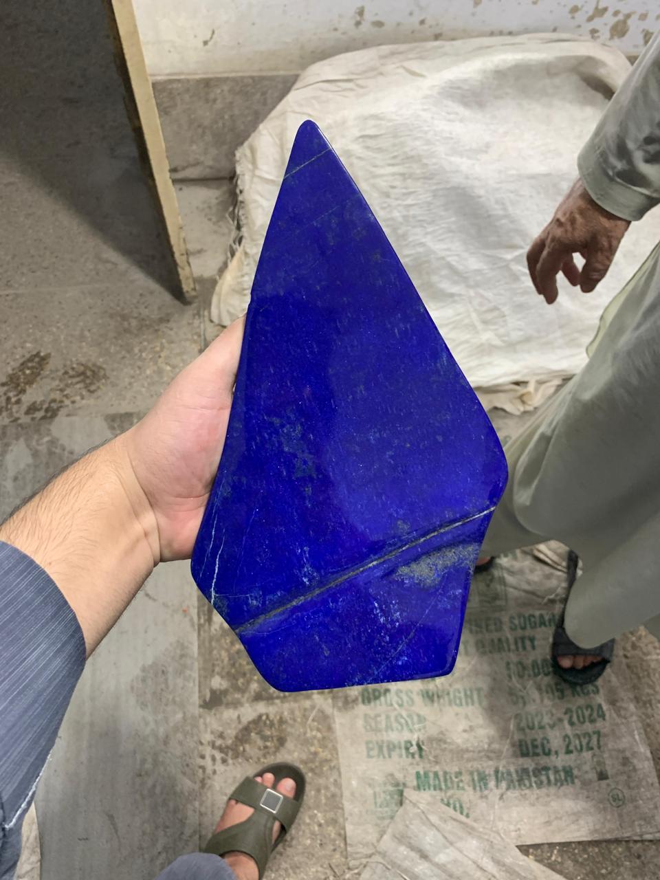 A hand holds a large, polished triangular piece of deep blue lapis lazuli stone in a workshop setting, with another person standing in the background near a white sack on a concrete floor.