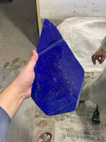 A hand holding a large, irregularly shaped dark blue stone slab, likely lapis lazuli, in a warehouse or workshop setting with another person partially visible in the background.