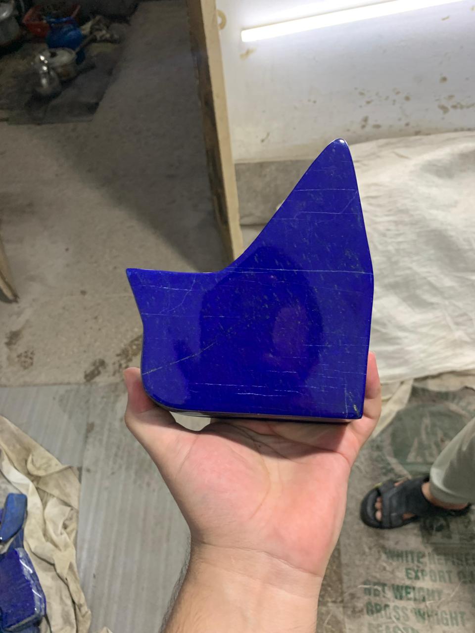 A hand holding a polished blue stone or crystal with an irregular triangular shape, photographed in a workshop or studio setting with fabric and tools visible in the background.