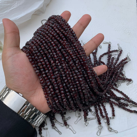 A hand holds a bundle of dark red faceted glass beads on strands with silver metal clasps, with more strands spread out on a white surface.