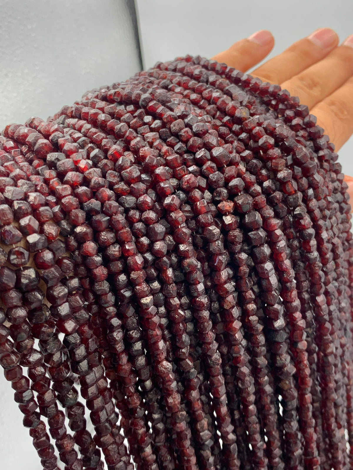 Multiple strands of faceted dark red gemstone beads held up by a hand against a light gray background.