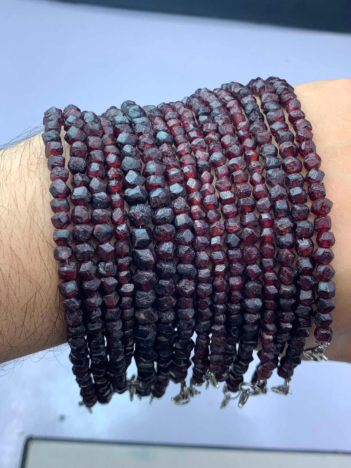 A close-up of a person's wrist wearing multiple strands of faceted dark red beads, likely gemstone beads, with small silver-toned charms at the ends.