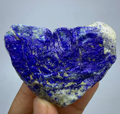 A raw, heart-shaped lapis lazuli crystal specimen held in a person's hand against a neutral light gray background. The stone features a deep blue and purple matrix with prominent gold pyrite veins and white calcite deposits.