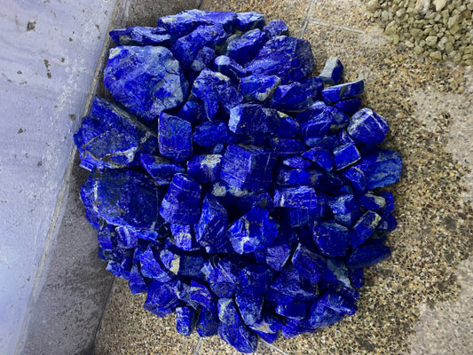A pile of raw, rough-cut cobalt blue lapis lazuli crystals resting on a concrete floor next to a gray wall.
