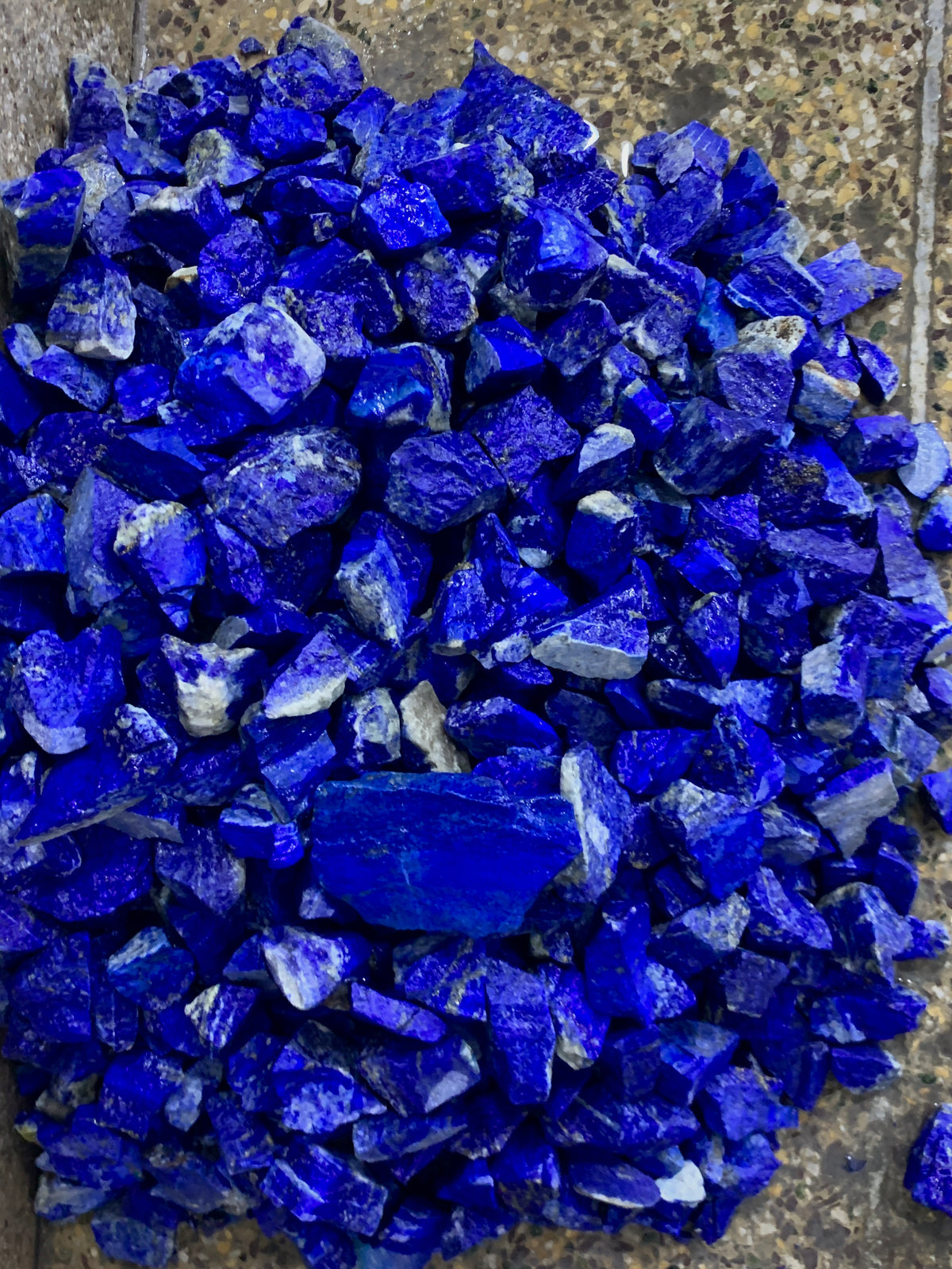 A pile of raw, rough-cut lapis lazuli crystal chips in vibrant deep blue, resting on a textured gray stone surface.