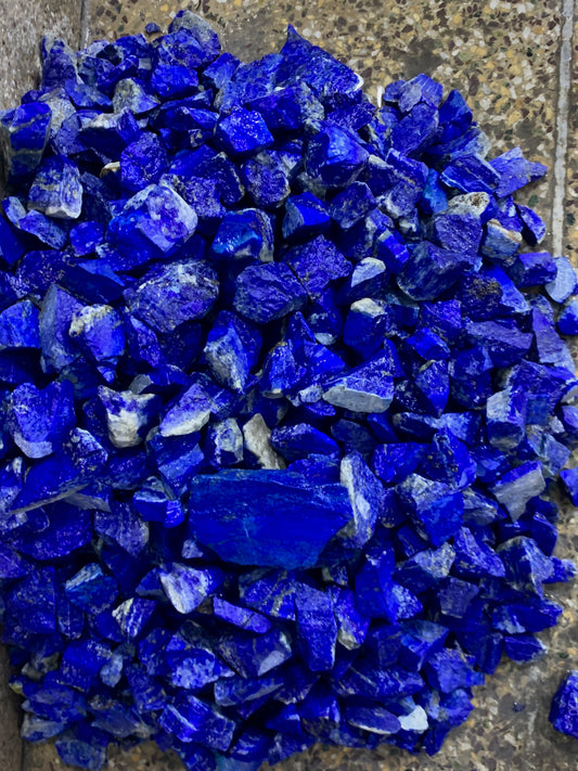A pile of raw, rough-cut lapis lazuli crystal chips in vibrant deep blue, resting on a textured gray stone surface.