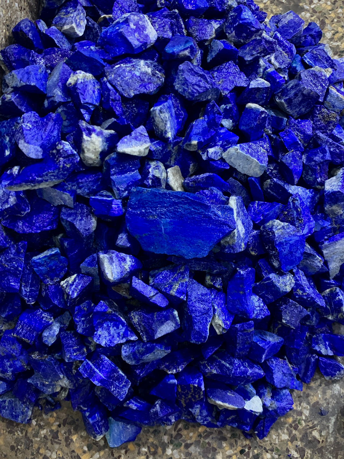 A pile of raw, rough-cut lapis lazuli crystal chips in vibrant deep blue, showing natural jagged edges and white veining, resting on a textured gray stone surface.