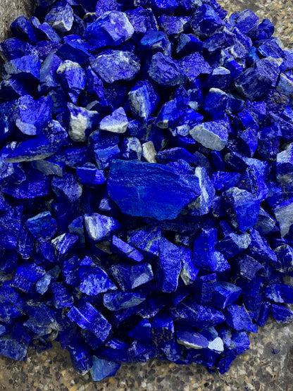 A pile of raw, rough-cut lapis lazuli crystal chips in vibrant deep blue, showing natural jagged edges and white veining, resting on a textured gray stone surface.