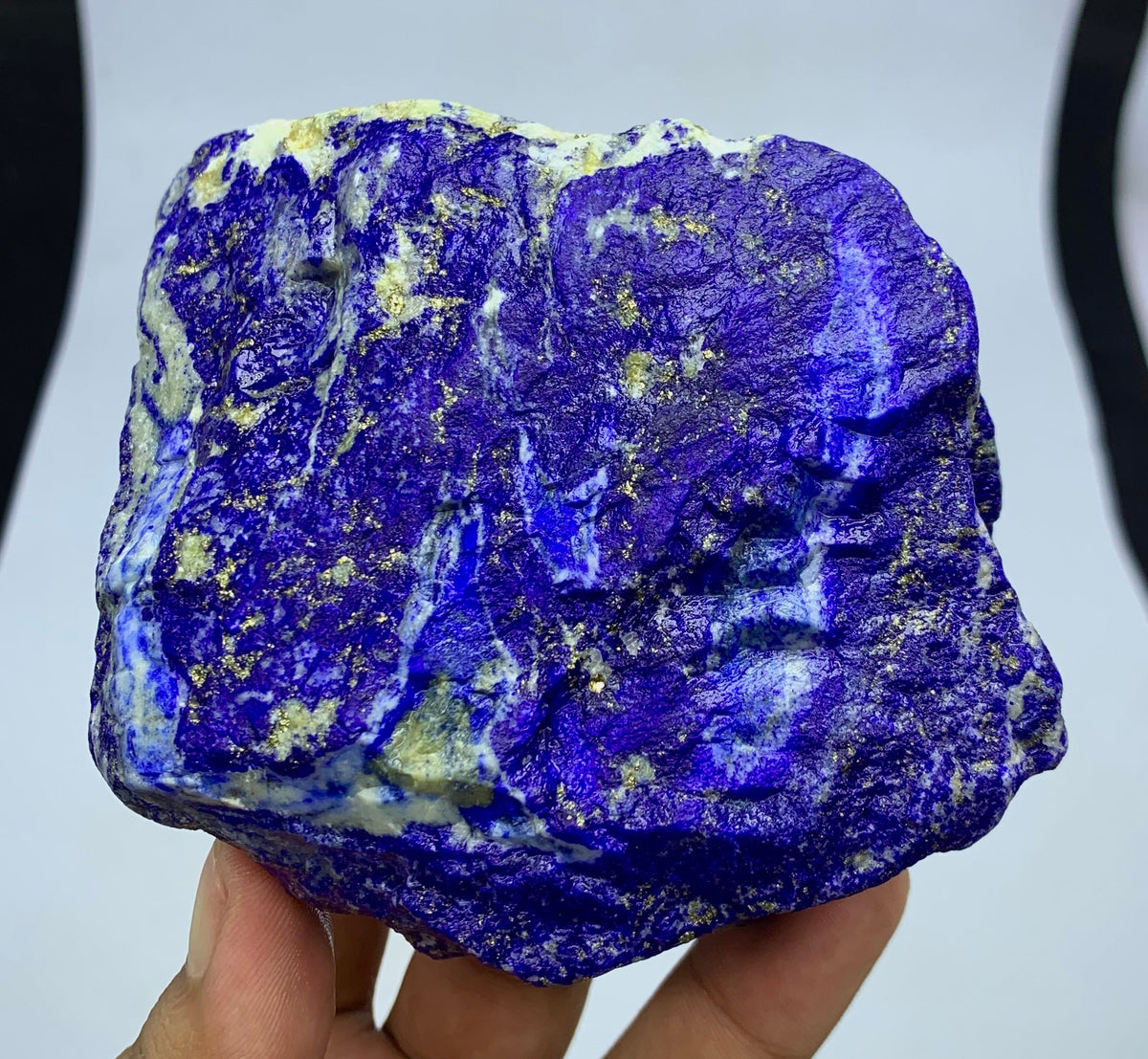 A hand holding a raw, rough-cut lapis lazuli crystal specimen showing deep blue-violet coloration with bright white and gold flecks, set against a black and white background.
Raw Lapis Lazuli specimen showing white Calcite streaks