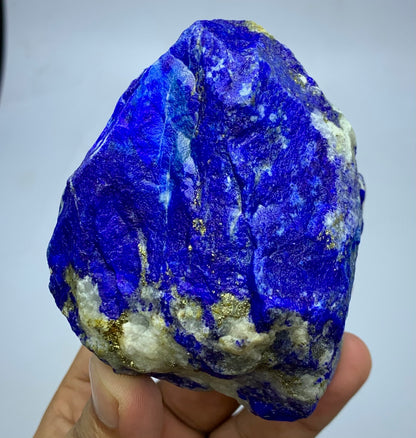 A raw blue lapis lazuli crystal specimen with gold pyrite and white calcite inclusions, held in a person's hand against a plain light background.