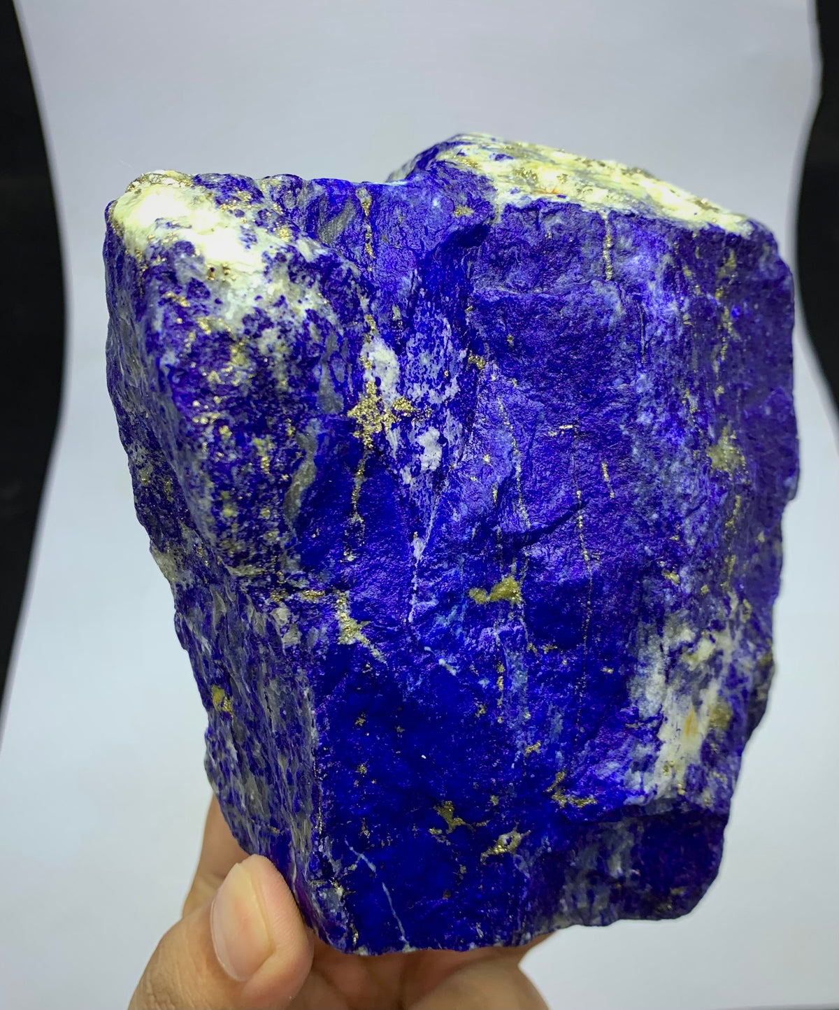 A raw blue lapis lazuli crystal specimen with prominent white and pale green matrix, held by a hand against a white background.