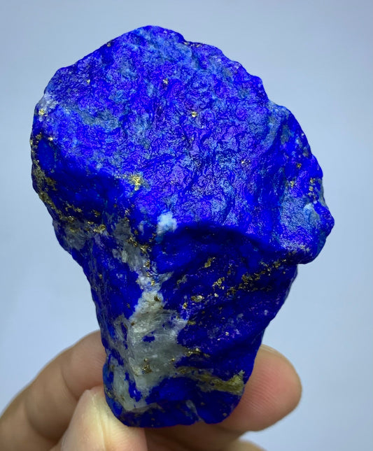 A raw, rough blue lapis lazuli crystal specimen with gold pyrite and white calcite inclusions, held by a hand against a light gray background.